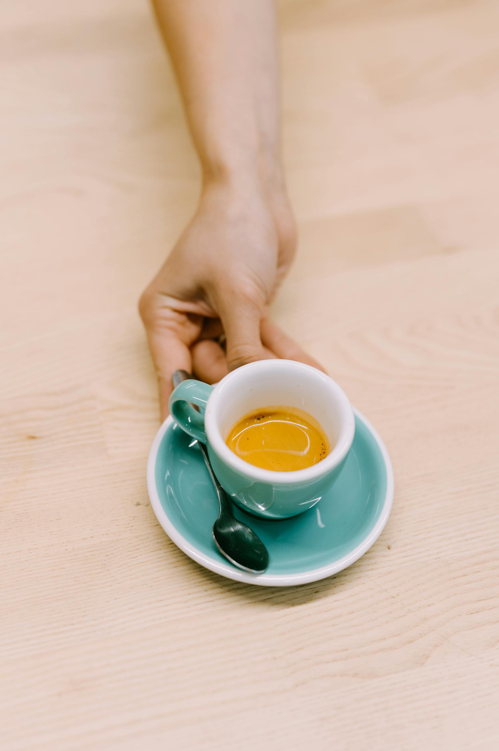 Close-up of a hand holding an espresso cup placed on a wooden table in a neutral cafe setting.
